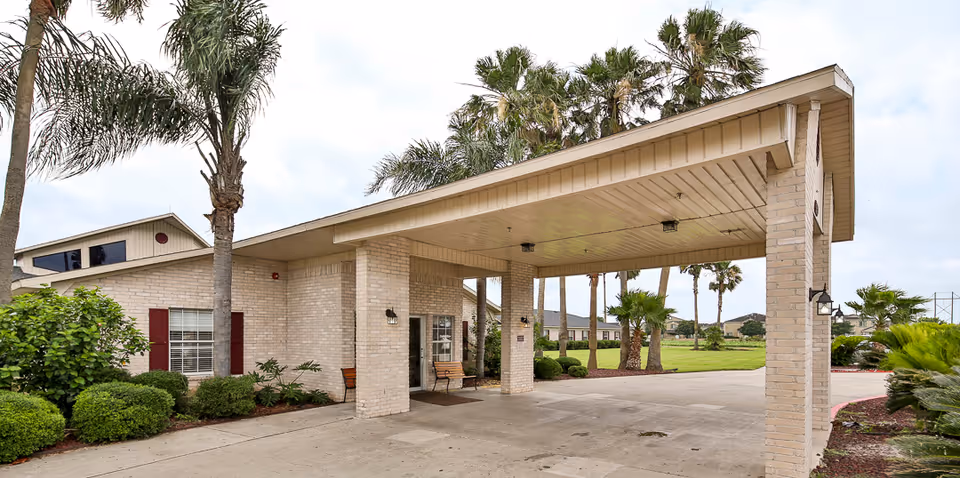Covered drive-through entrance with brick columns and a bench at the front of a nursing and rehabilitation building, surrounded by palm trees and landscaping.