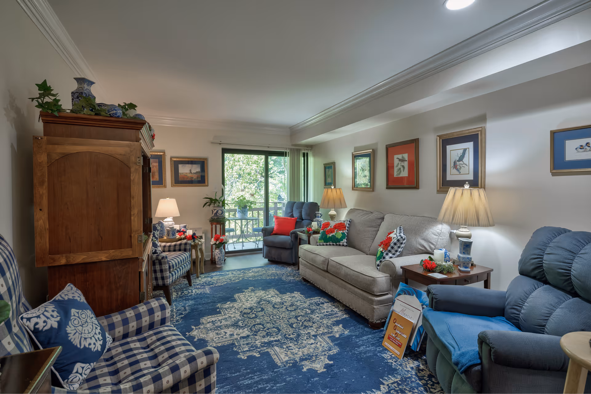A cozy living room with a blue patterned rug, a beige sofa with floral and checkered pillows, two blue armchairs, a wooden cabinet, and several framed pictures on the walls. There are two table lamps on side tables, and a sliding glass door leading to a balcony with greenery outside.