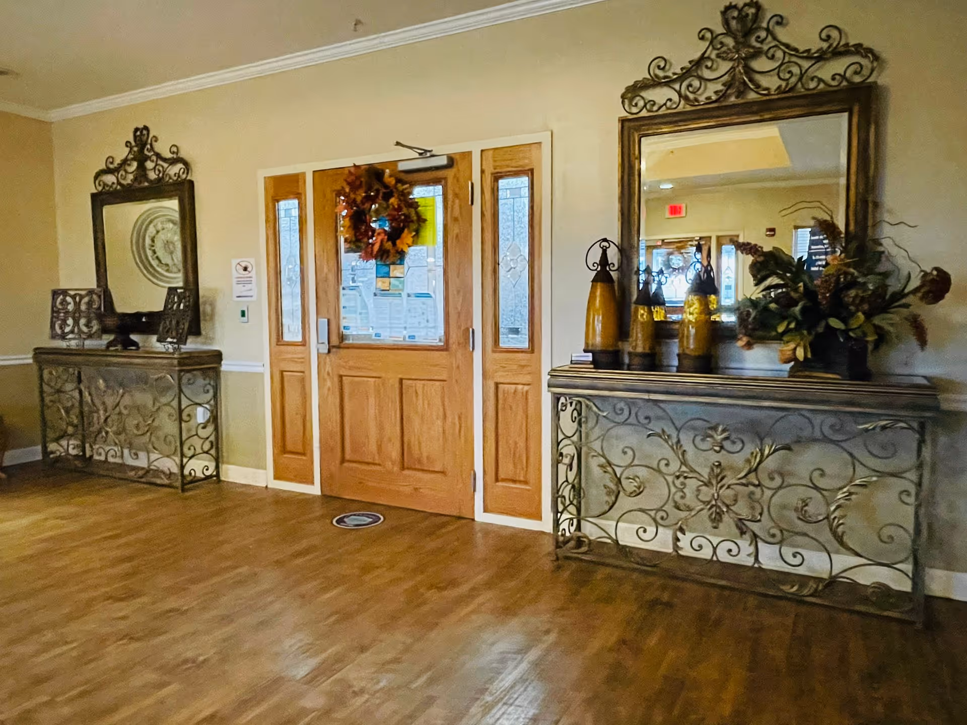 Interior view of an entrance area with double wooden doors decorated with a fall wreath. On either side of the doors are ornate metal and wood console tables with large decorative mirrors above them. The right table has three yellow decorative vases and a floral arrangement. The floor is wooden and the walls are painted beige.