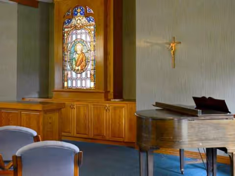 Wood-paneled chapel area with a stained glass window, a grand piano, and blue chairs.