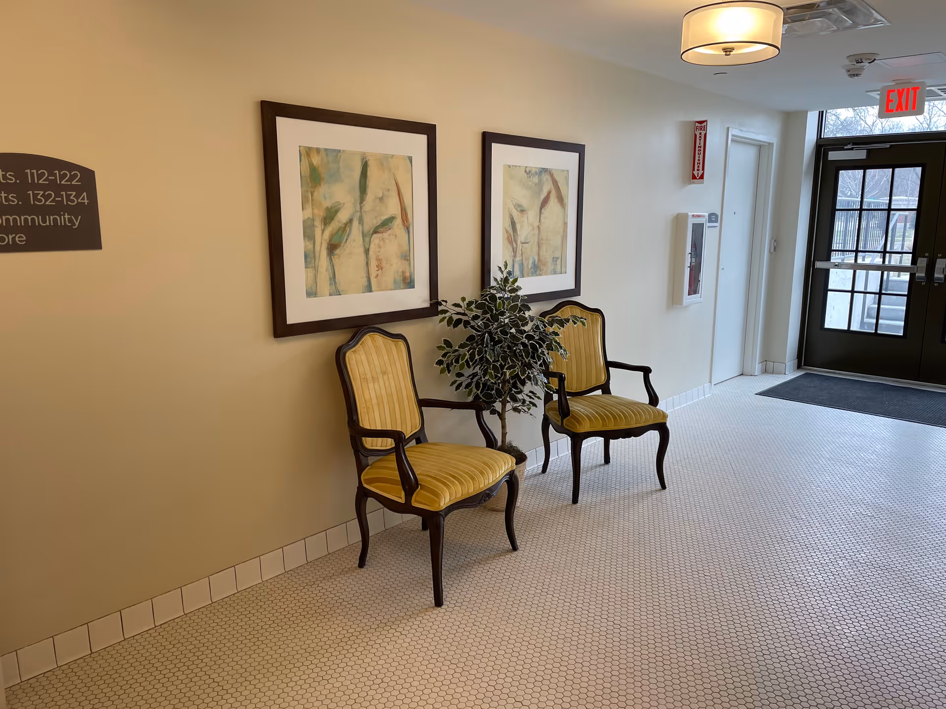 A hallway area in Bardwell Residences with two yellow upholstered wooden chairs placed against a beige wall. Above the chairs are two framed abstract paintings. A potted plant is situated between the chairs. The floor is covered with small white hexagonal tiles. A glass door with an exit sign above it is visible at the end of the hallway, along with a fire extinguisher cabinet and a fire alarm on the wall.