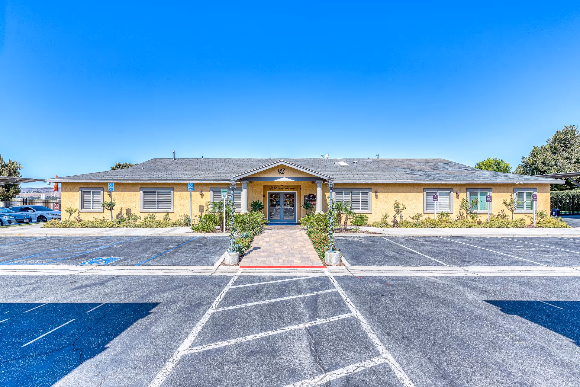Front exterior view of a single-story yellow building with a gray roof under a clear blue sky. There is a paved walkway leading to double glass doors at the entrance, flanked by plants and small bushes. Several parking spaces, including handicapped spots, are visible in front of the building.
