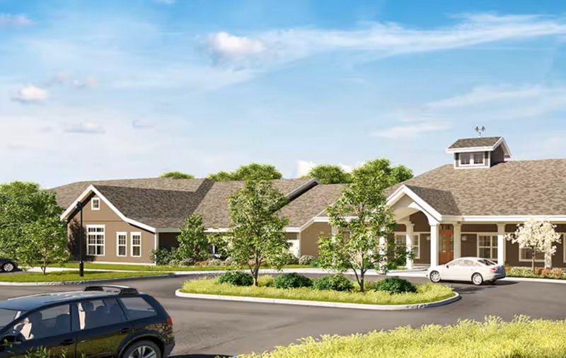 Exterior view of a single-story senior living facility building with a circular driveway, landscaped greenery, and parked cars under a blue sky with some clouds.