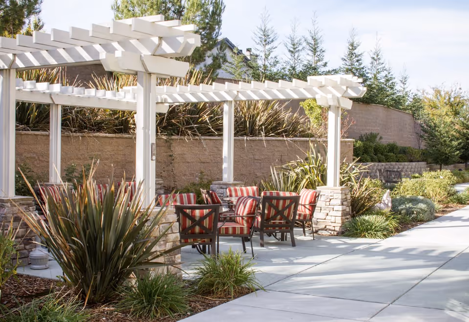 Outdoor seating area with a white pergola, several cushioned chairs with red and white striped upholstery, surrounded by plants and greenery along a paved walkway.