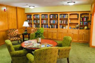 A cozy library or reading room with a round wooden table surrounded by four green upholstered chairs. There are bookshelves filled with books along the back wall, a wooden side table with a lamp, and two patterned armchairs in the corner. The room has green carpet and wood-paneled walls.