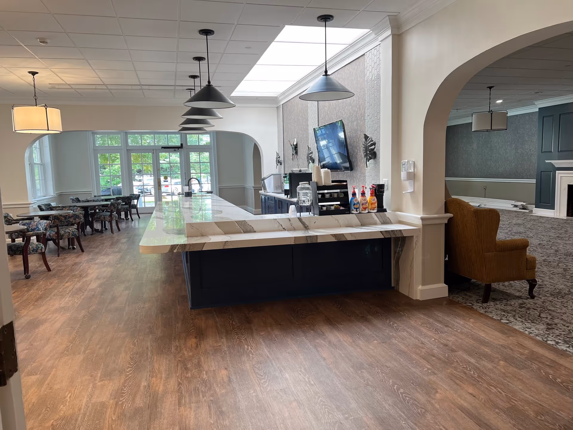 Interior view of a senior living facility showing a long marble countertop with pendant lights above it, a beverage station with coffee and syrup dispensers, and a seating area with tables and floral upholstered chairs near large windows. Adjacent to this area is a lounge with a carpeted floor, an armchair, and a fireplace.
