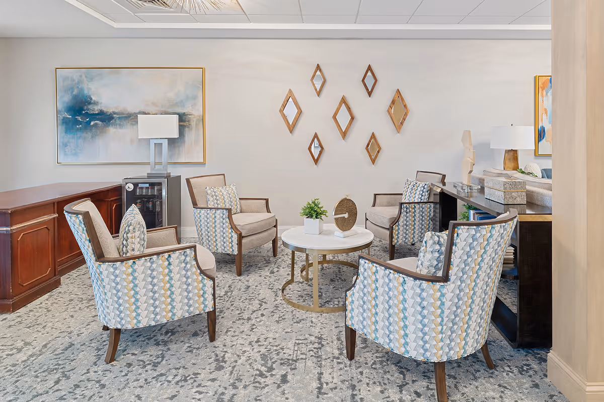 A cozy seating area in a senior living facility with four patterned armchairs arranged around a round white marble coffee table. The room features a light gray carpet with a subtle pattern, a wooden sideboard with a lamp and a mini fridge, abstract wall art, and decorative diamond-shaped mirrors on the wall.
