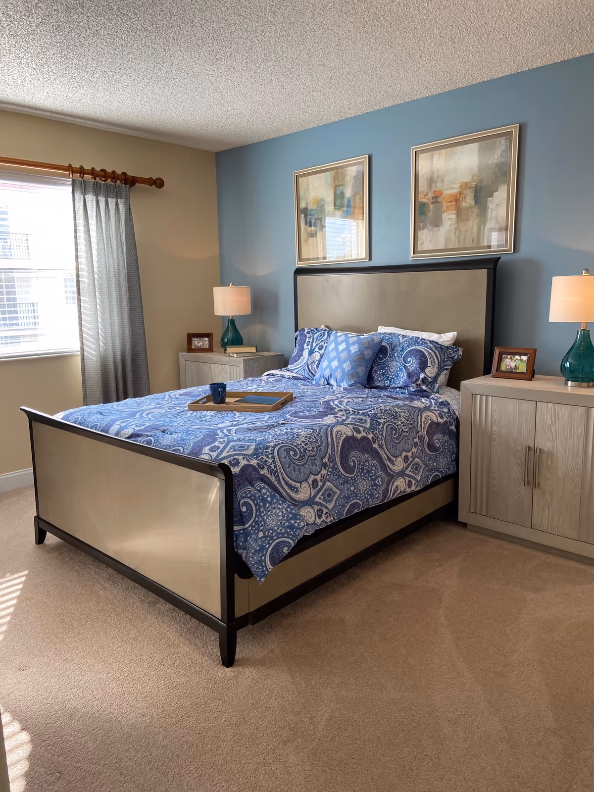Well-appointed bedroom with a bed dressed in blue patterned bedding, two nightstands with lamps, framed artwork above the headboard, and a window with curtains.