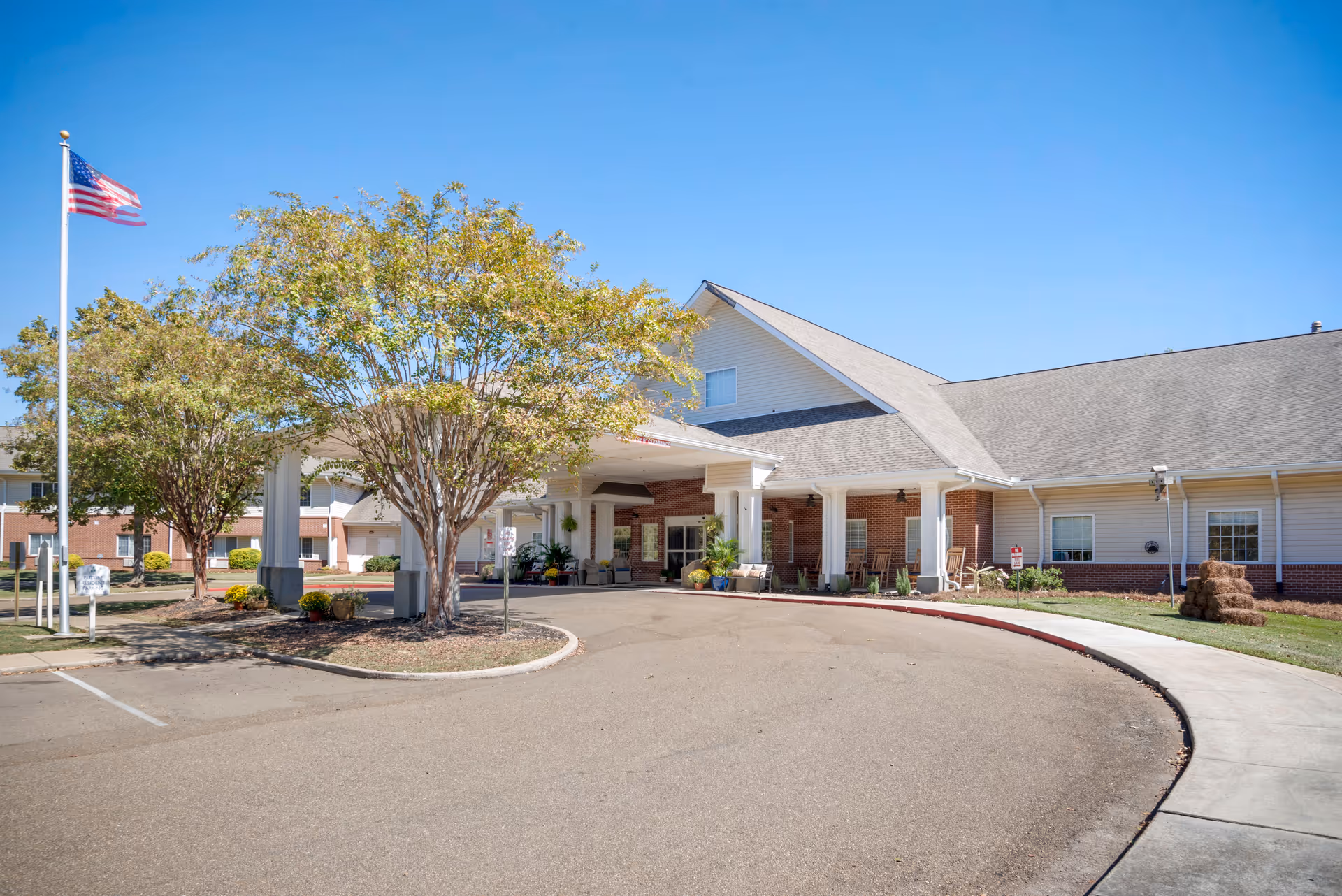 Exterior view of Brookdale Clinton senior living facility showing the main entrance with a covered drop-off area, trees, an American flag on a flagpole, and a clear blue sky.