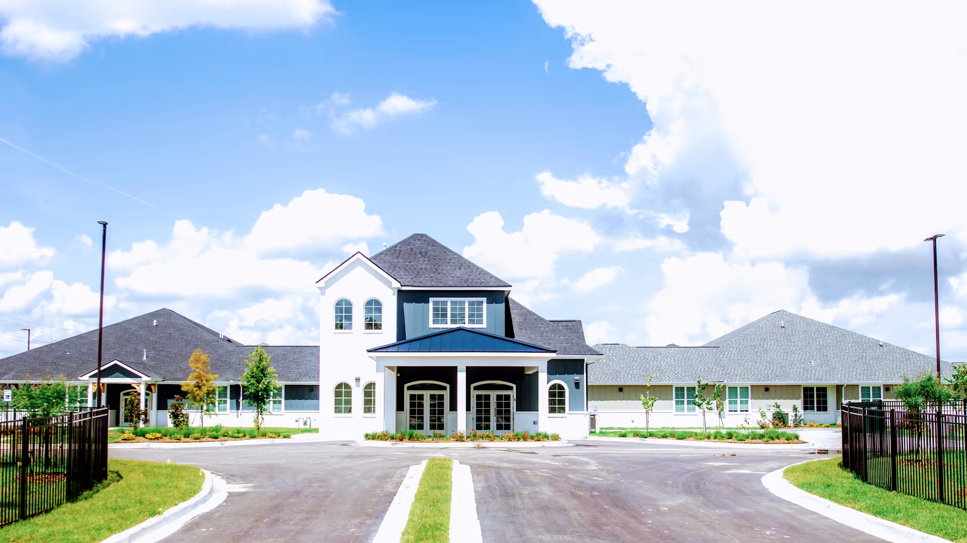 Front exterior view of Sage Oak of Lake Charles facility under a partly cloudy blue sky, featuring a large driveway, manicured lawn, and a building with multiple roof peaks and arched windows.