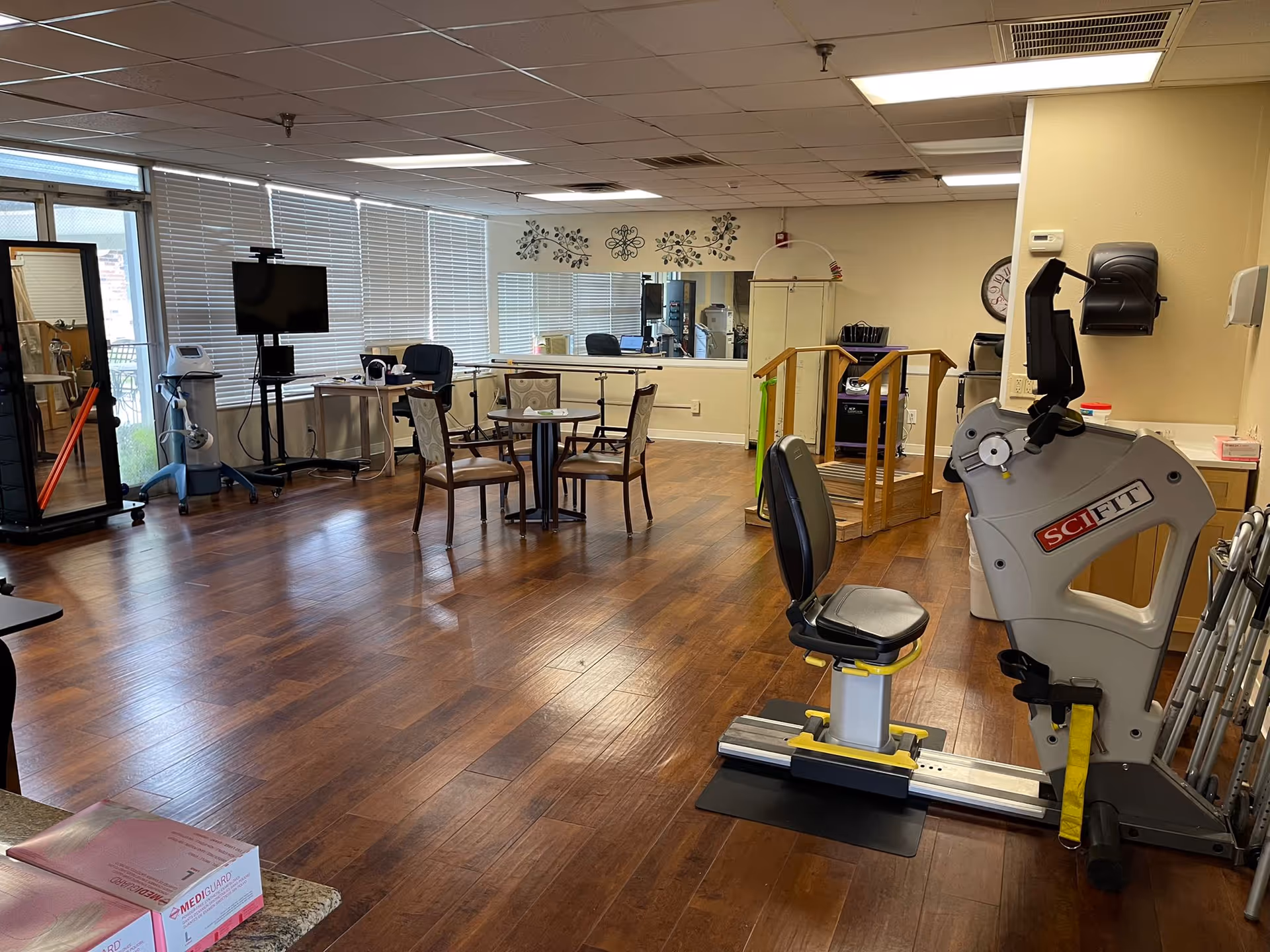 A spacious senior activity/therapy room with exercise equipment, a recumbent exercise machine in the foreground, tables and chairs, a TV and large windows.