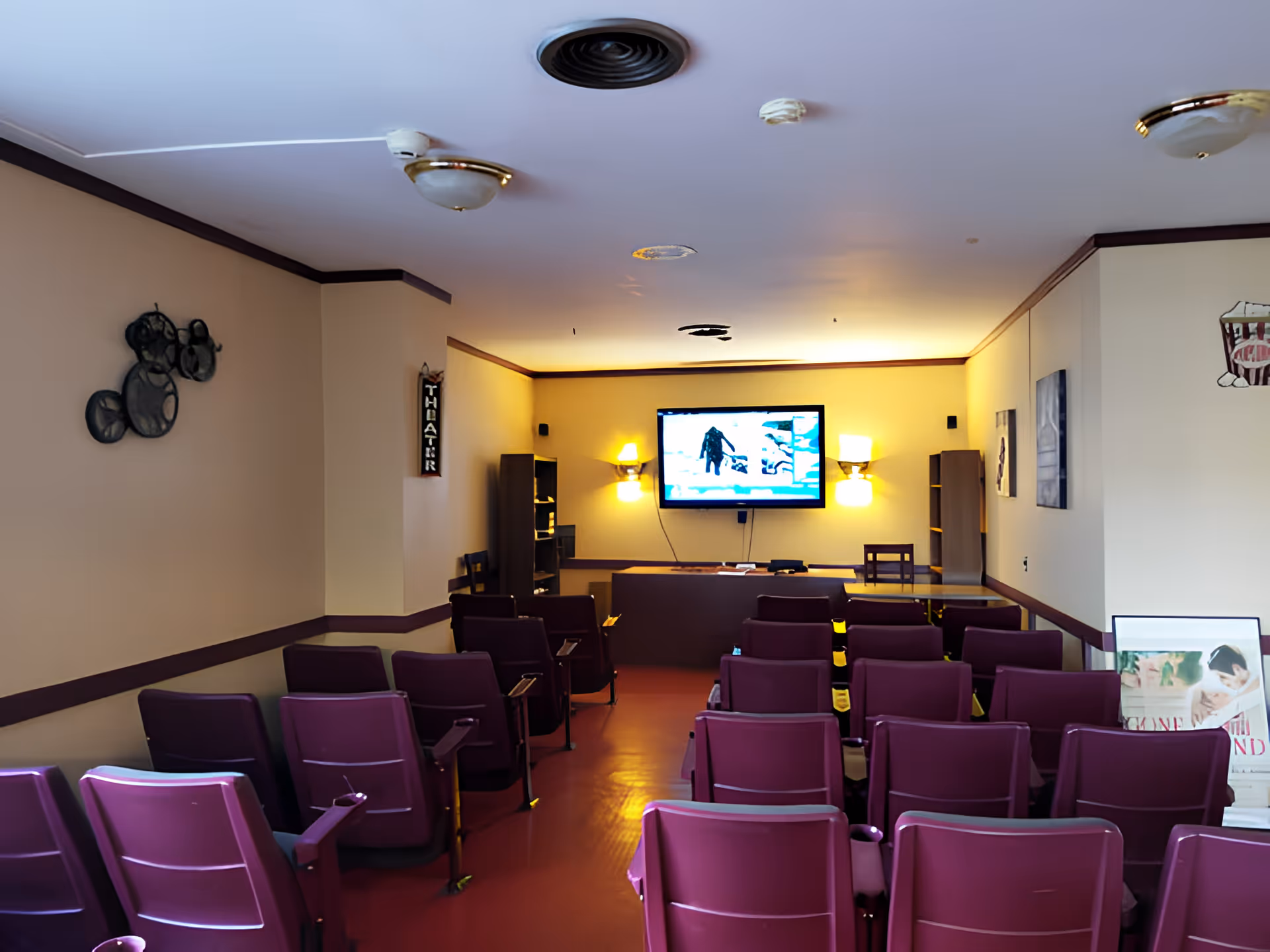 A small theater room with rows of purple chairs facing a wall-mounted flat screen TV. The room has beige walls with dark trim, wall sconces providing warm lighting, and decorative wall art. There is a sign on the left wall that reads 'THEATER'.