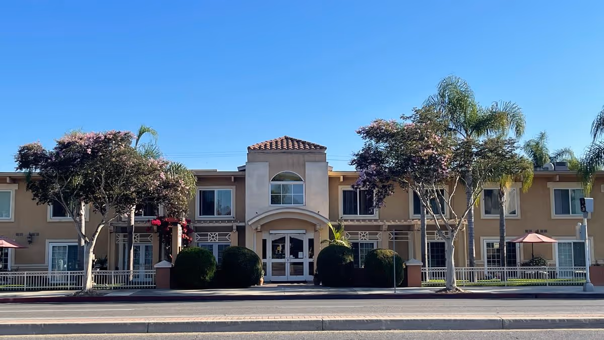 Front exterior of a two-story beige senior living building with a central arched entrance, palm trees, and landscaping.