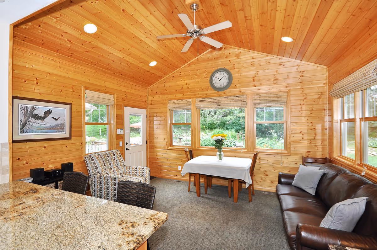 Interior view of a cozy room with wooden walls and ceiling, featuring a ceiling fan, a small dining table with a white tablecloth and a vase of flowers, a brown leather sofa with gray pillows, a patterned armchair, and large windows showing greenery outside.