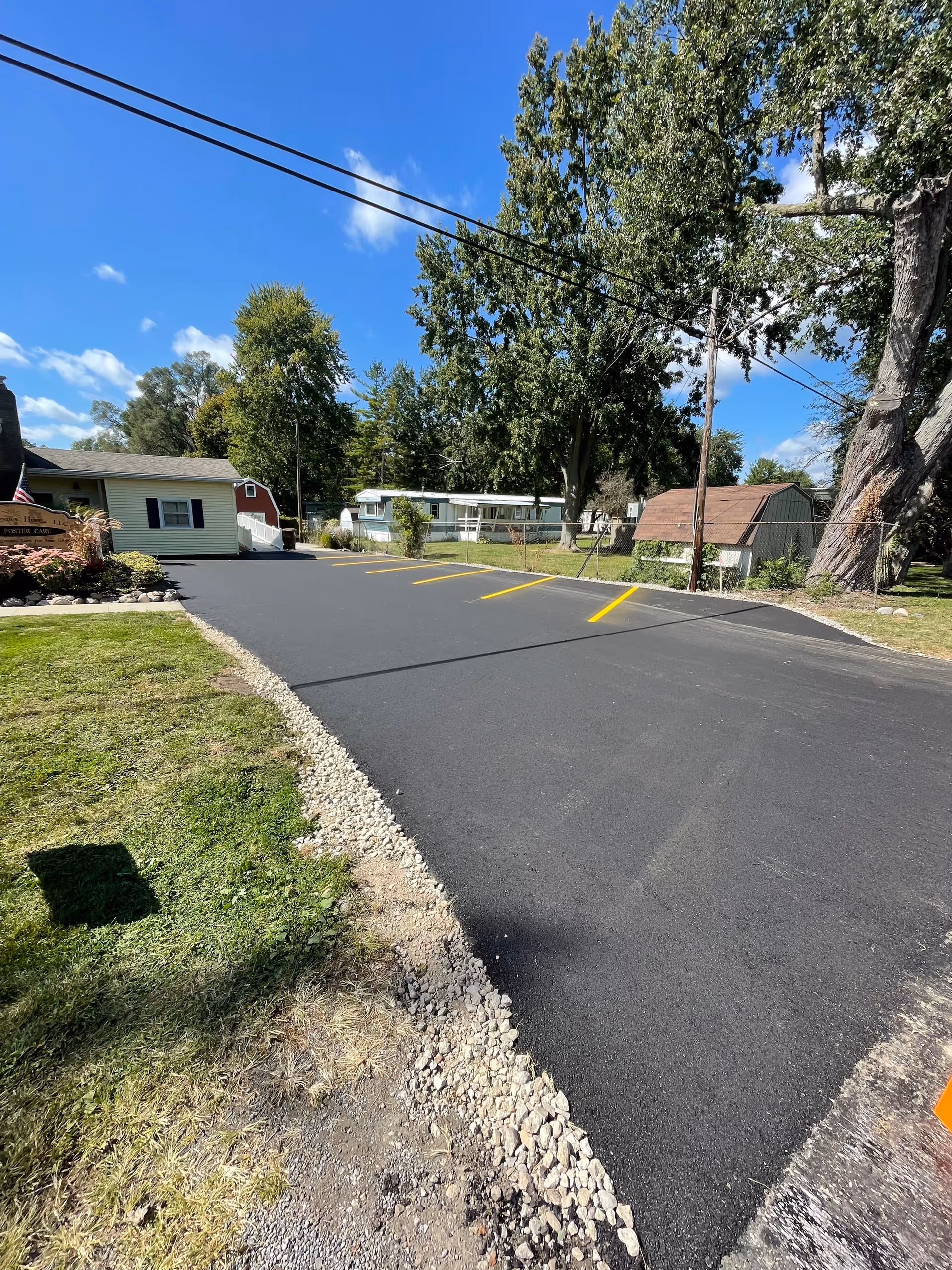 Newly paved parking lot with yellow parking lines in front of a small single-story building and large trees under a blue sky.