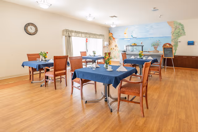 A bright dining room with wooden floors and several tables covered with blue tablecloths, each set with white napkins, cups, and small flower arrangements. The room has a large window with curtains, a wall clock, and a colorful mural depicting a seaside scene with sailboats on the far wall. There is also a small buffet area with a chalkboard menu and a cabinet along the wall.