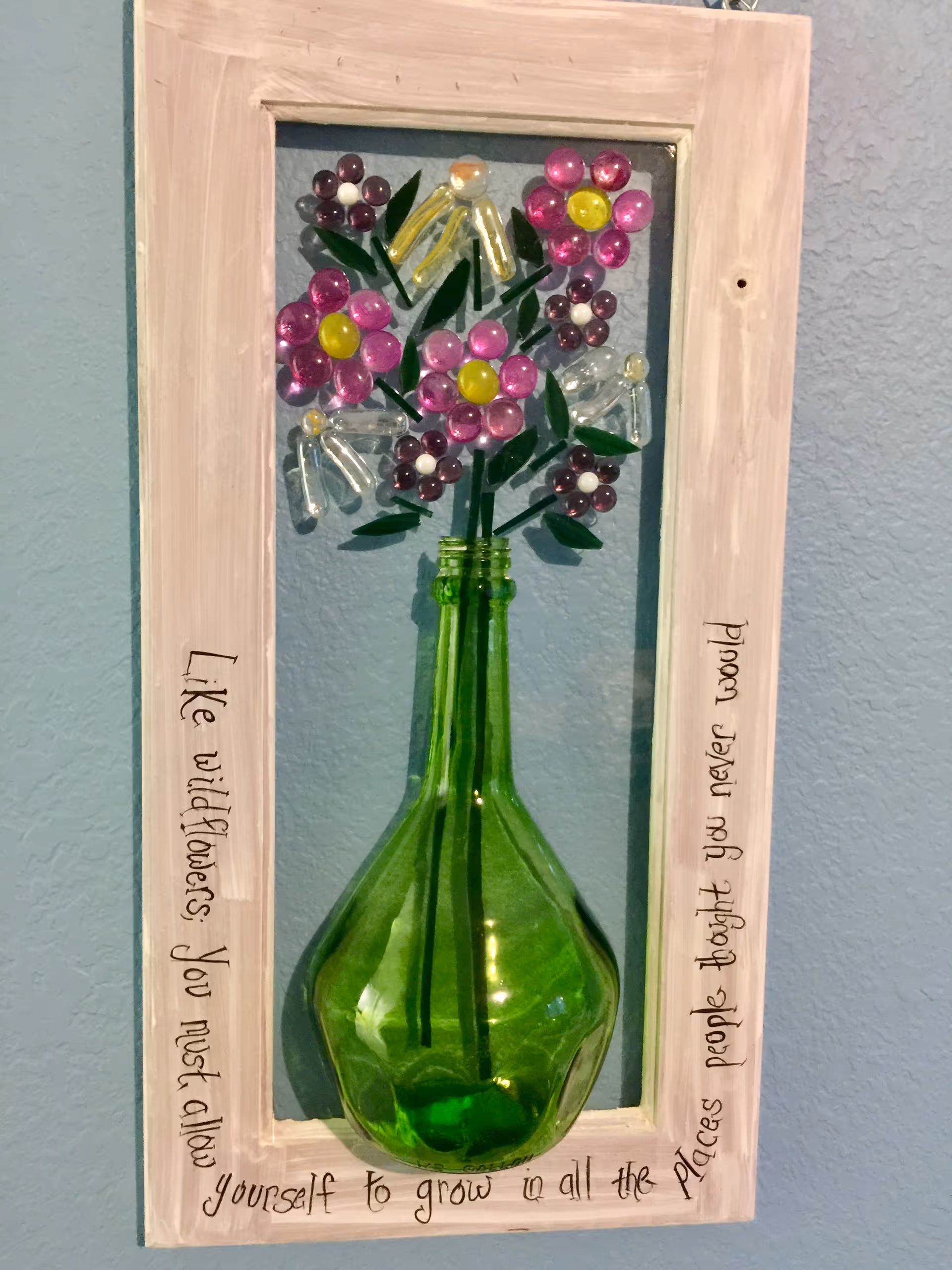 Framed decorative glass flower arrangement in a green bottle mounted on a blue wall.