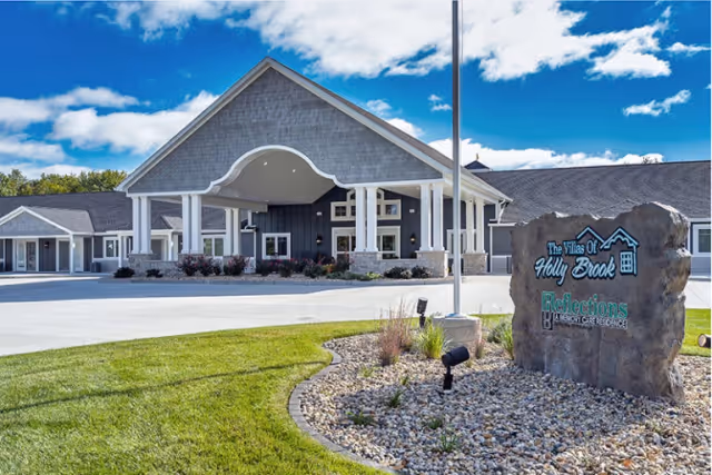 Front entrance of a single-story assisted living facility with a covered porte-cochère and a stone sign in a landscaped rock bed.
