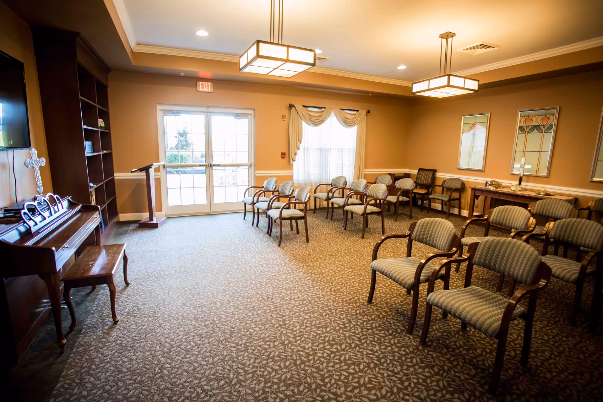 A well-lit room with rows of striped upholstered chairs arranged facing a wooden podium near glass double doors. The room has beige walls, carpeted floor with a leaf pattern, a piano with a cross on it, and framed artwork on the walls. Ceiling lights provide warm illumination.