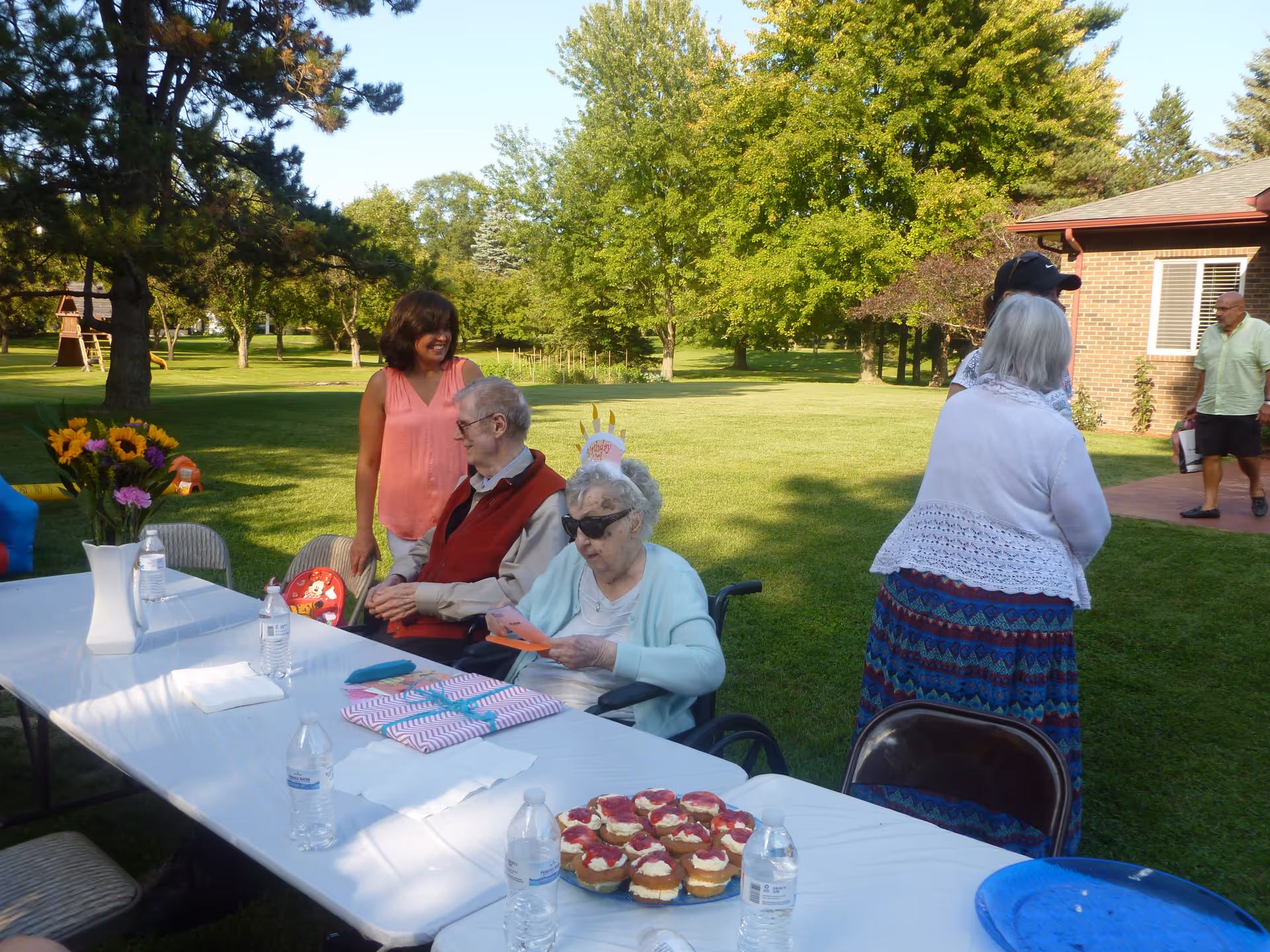 A group of elderly people and a caregiver gathered outdoors at a senior living facility. They are sitting and standing around a long table covered with a white tablecloth, which has water bottles, a vase with flowers, wrapped gifts, and a plate of cupcakes. The setting is a green grassy area with trees and a brick building in the background, under clear daylight.
