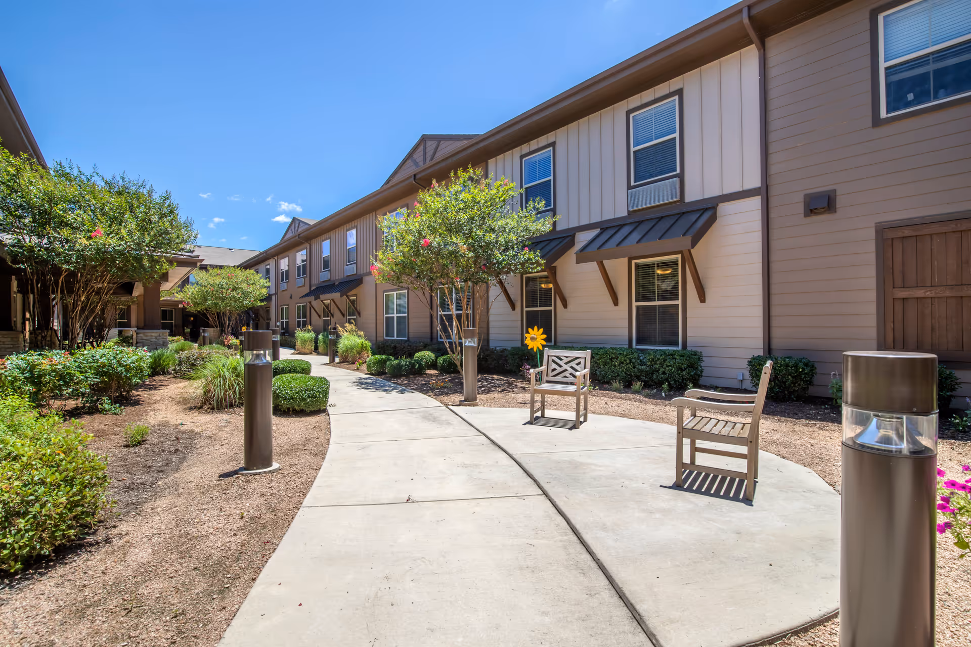 Outdoor courtyard area of Landon Ridge Alamo Ranch Independent Living featuring a curved concrete walkway, two wooden chairs, small trees, bushes, and a two-story building with multiple windows and awnings under a clear blue sky.