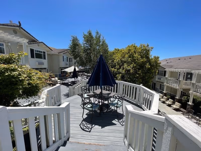 Outdoor patio area at Fremont Hills with a wooden deck, metal table and chairs with a closed blue umbrella in the center. Surrounding the deck are trees and shrubs, with multi-story residential buildings in the background under a clear blue sky.