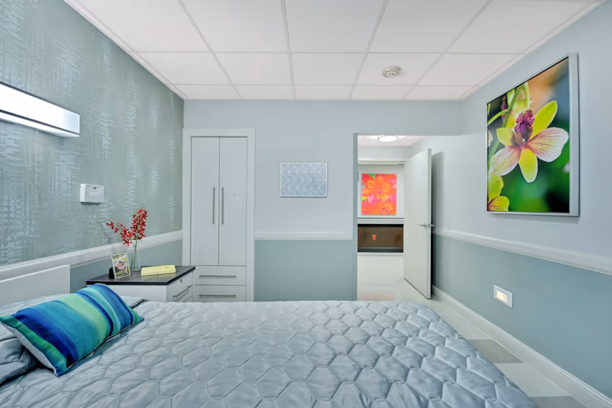 A neatly made bed with a gray quilt and a blue-green striped pillow in a nursing center bedroom. The room features light blue walls with a white trim, a small nightstand with a vase of red flowers and a framed photo, a closed white wardrobe, and colorful floral artwork on the walls. An open door reveals a hallway with another bright floral picture.