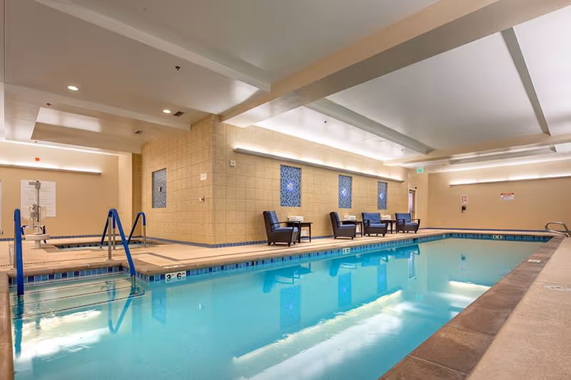 Indoor swimming pool area with clear blue water, beige tiled walls, and ceiling lights. There are several dark chairs and small tables along the far wall, and a hot tub is visible in the background to the left.