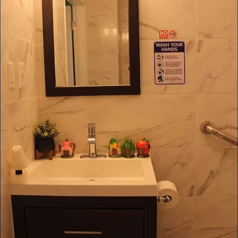 Small bathroom sink with a mirror above, faucet, grab bar, a 'Wash Your Hands' sign on the tiled wall, and decorative figurines on the counter.