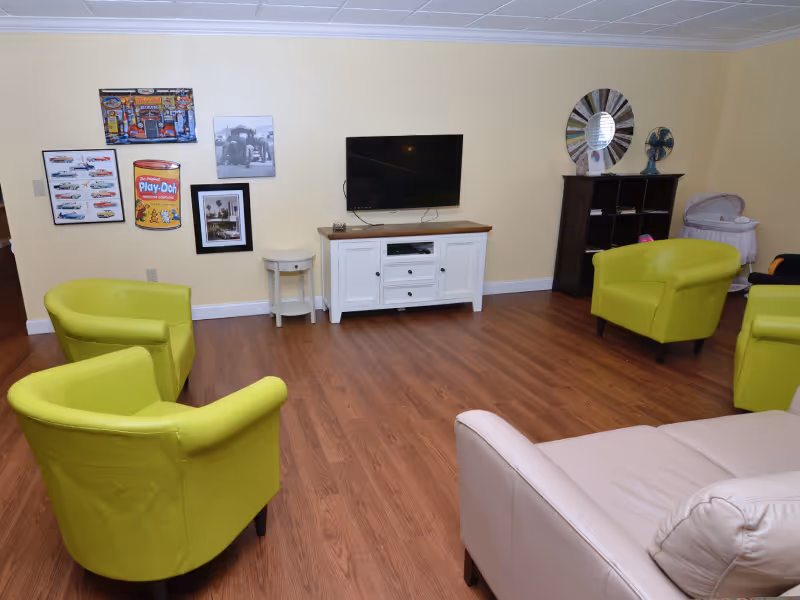 A living room with four lime green armchairs and a beige sofa arranged around a wooden floor. A flat-screen TV is mounted on a beige wall above a white wooden cabinet. The walls are decorated with framed pictures and a large round mirror above a dark wooden shelving unit. A small white side table and a baby bassinet are also visible.