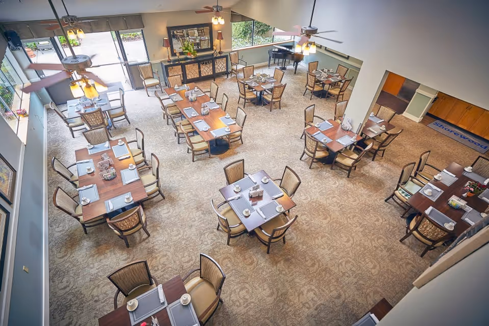 A spacious dining room in a senior living facility with multiple wooden tables and cushioned chairs arranged neatly. Each table is set with placemats, cups, and napkins. The room has large windows allowing natural light, ceiling fans, and a piano in the corner. A mirror and decorative plants are visible on the far wall.