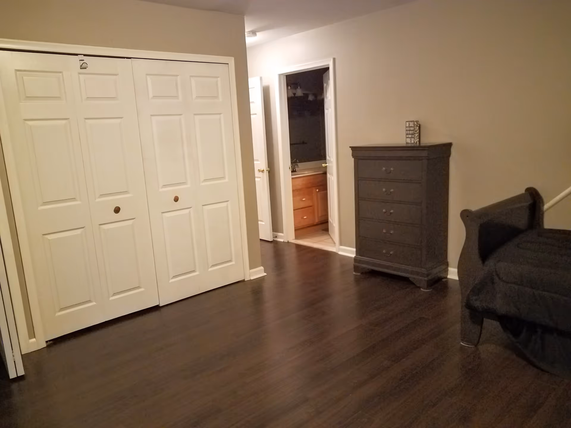 Interior view of a room with dark wooden flooring, beige walls, a dark wooden dresser with five drawers, a dark wooden bed frame with a black mattress, and white double closet doors. An open door reveals a bathroom with a wooden vanity and a sink.