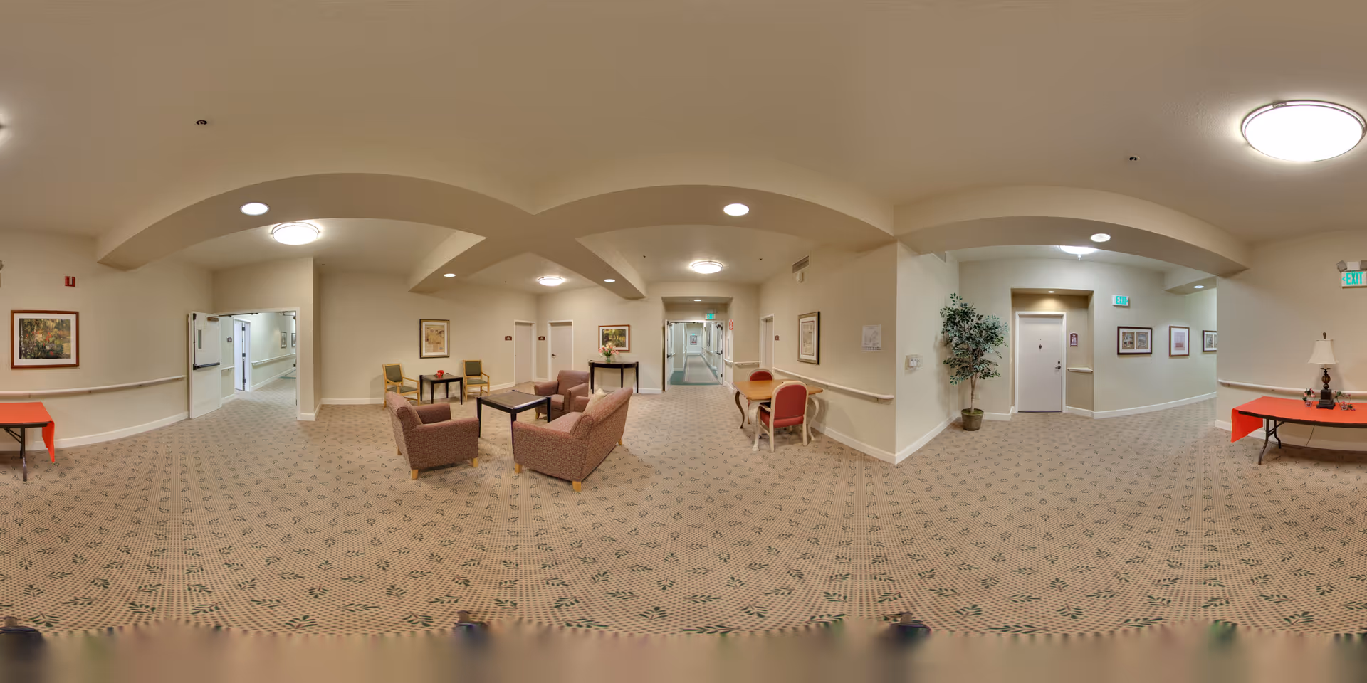 A spacious senior living facility common area with beige walls and patterned carpet. The room features several upholstered armchairs and a sofa arranged around a coffee table in the center. There are additional chairs and small tables along the walls, framed artwork, potted plants, and ceiling lights providing illumination. Multiple doorways and hallways lead to other parts of the facility.