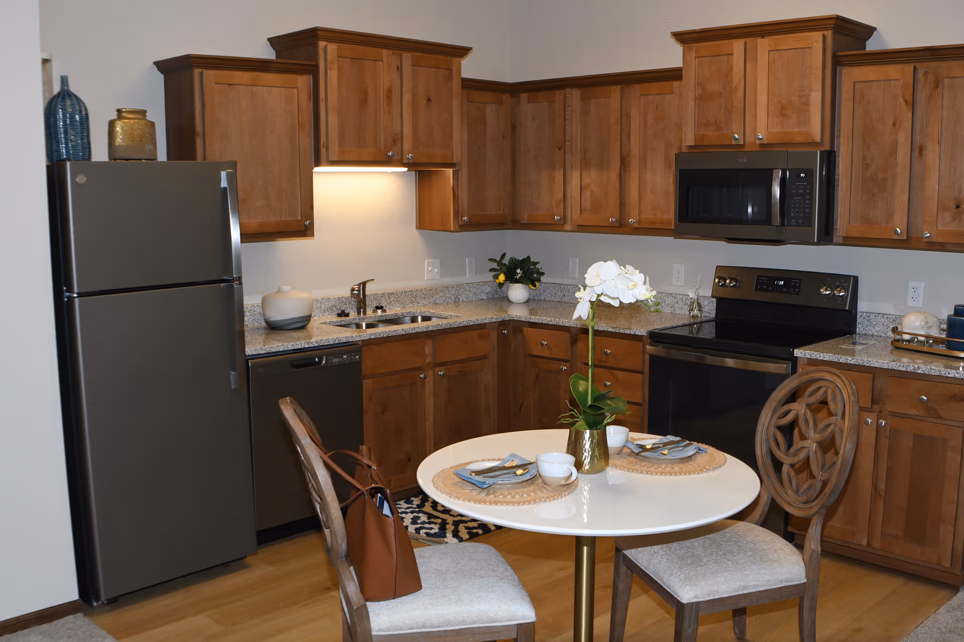 Modern kitchenette with wooden cabinets, stainless-steel appliances, and a small round dining table set for two.