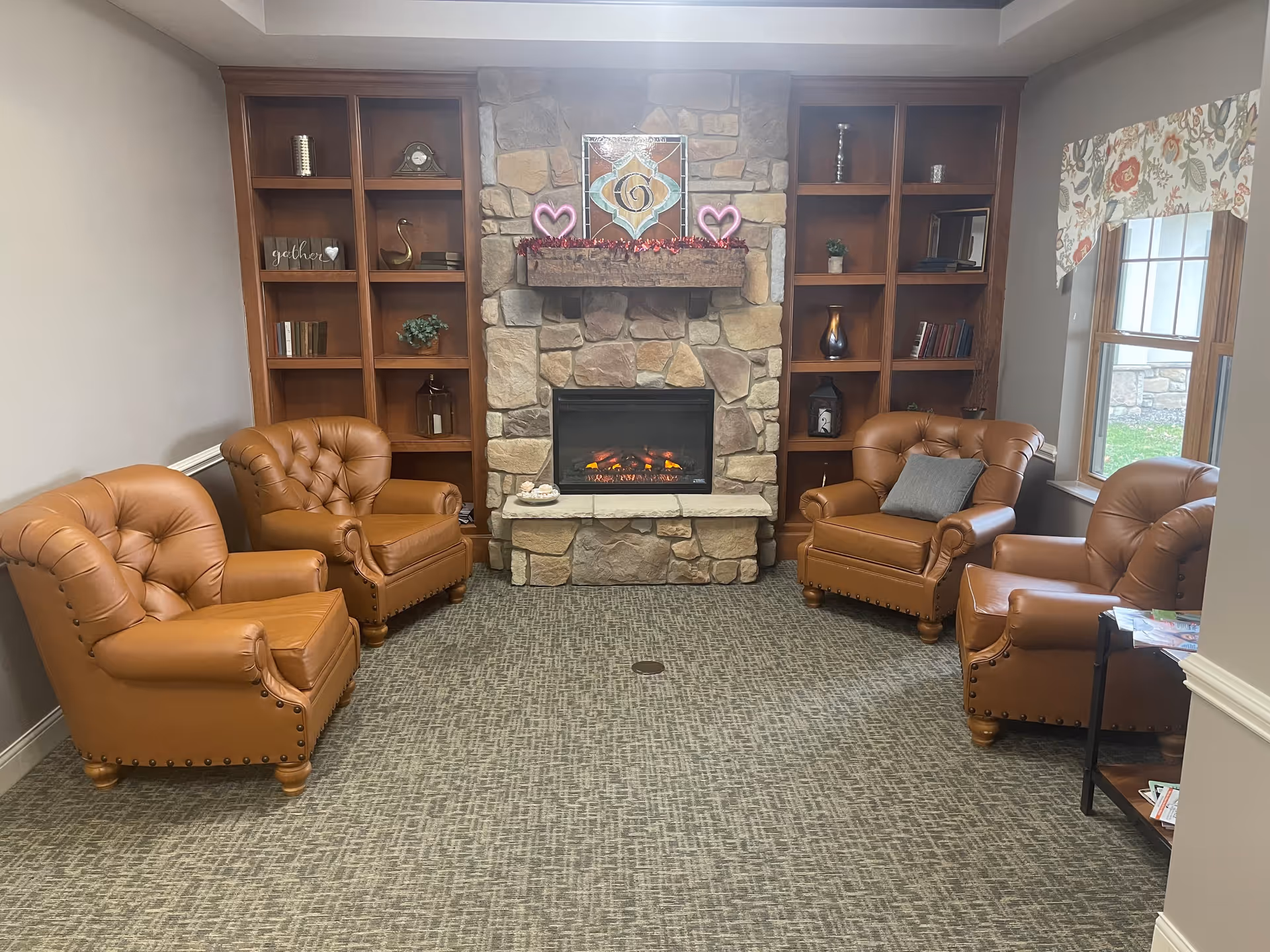 A cozy living room area with four brown leather armchairs arranged around a stone fireplace with a wooden mantel. The mantel is decorated with two pink heart-shaped lights and a stained glass piece with the letter 'G'. Built-in wooden shelves flank the fireplace, holding decorative items and books. A window with floral valance is on the right side, letting in natural light.