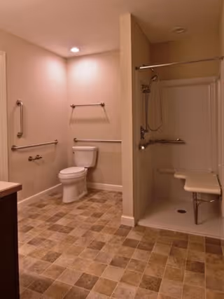 A spacious bathroom with beige walls and tiled floor. The room features a white toilet with grab bars on the walls around it, and an open shower area with a fold-down seat and a handheld showerhead. The lighting is recessed in the ceiling.