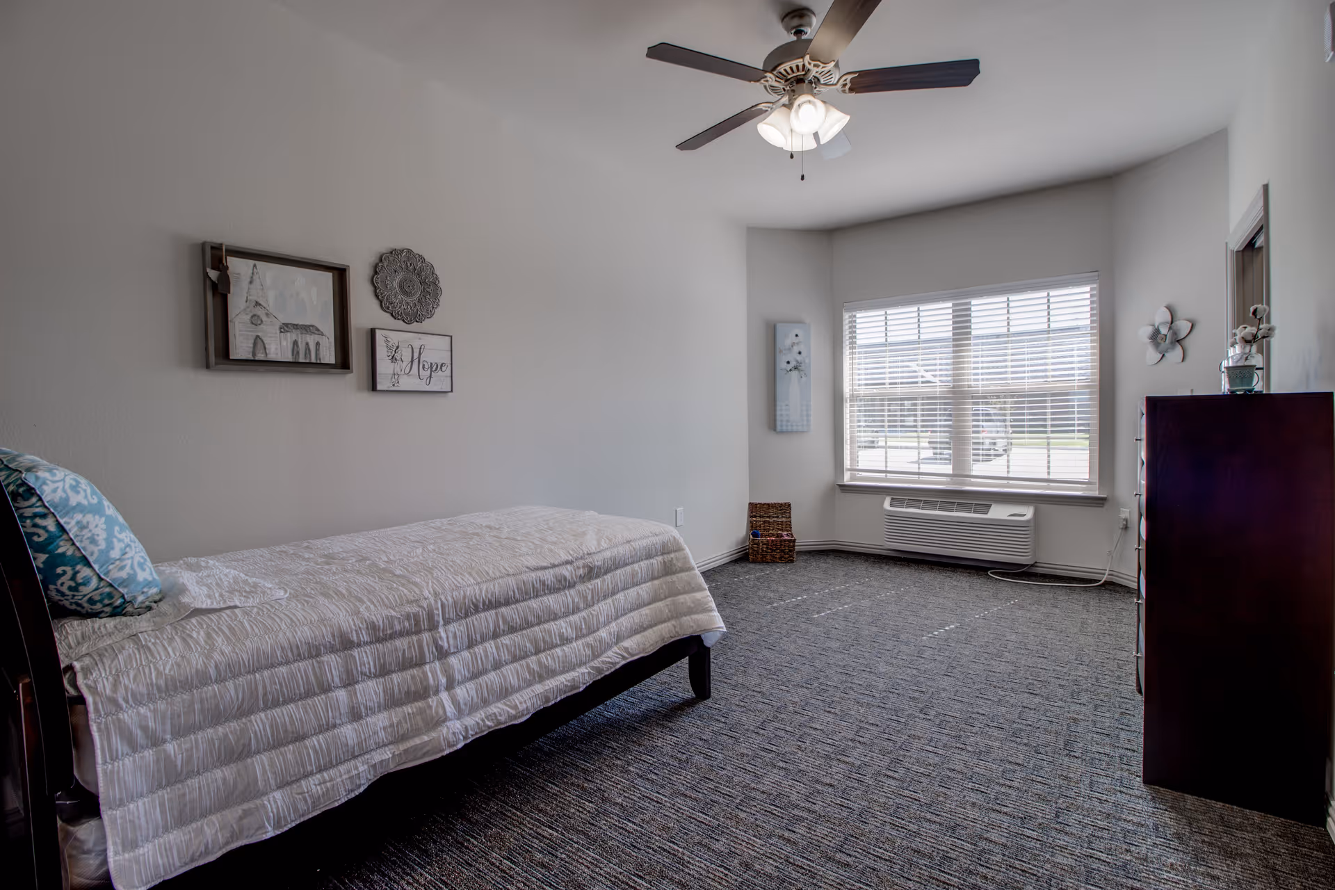Bright single-occupancy bedroom with a twin bed, ceiling fan, dresser, wall decorations and a large window with blinds.