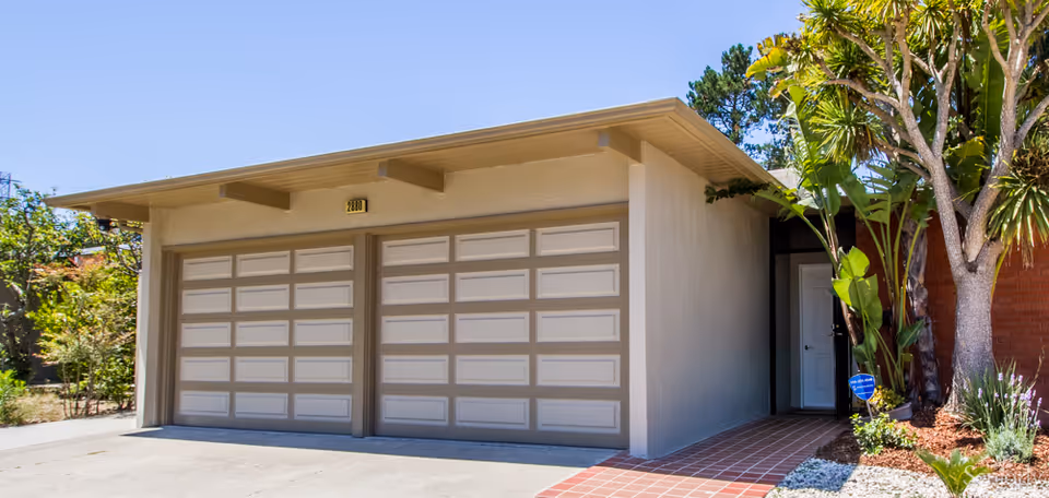 Front exterior of a small building featuring two closed garage doors, a tiled entryway, and surrounding landscaping.
