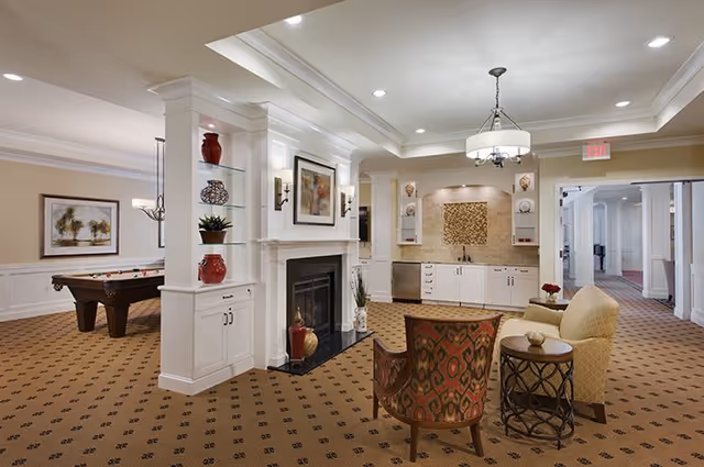 A spacious and well-lit common area in an assisted living facility featuring a fireplace with decorative shelves, comfortable seating including an armchair and a sofa, a small round table, and a kitchenette area with cabinets and a sink. In the background, there is a pool table and framed artwork on the walls. The room has a patterned carpet and recessed lighting.