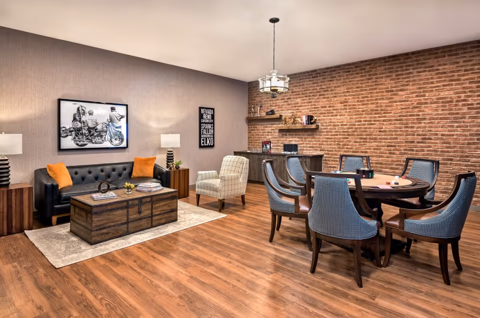 A cozy common area with a black leather sofa adorned with orange pillows, a wooden coffee table on a beige rug, two side tables with lamps, a single armchair, and a round table with six upholstered chairs. The room features a brick accent wall with floating shelves and a cabinet, wooden flooring, and a modern ceiling light fixture.