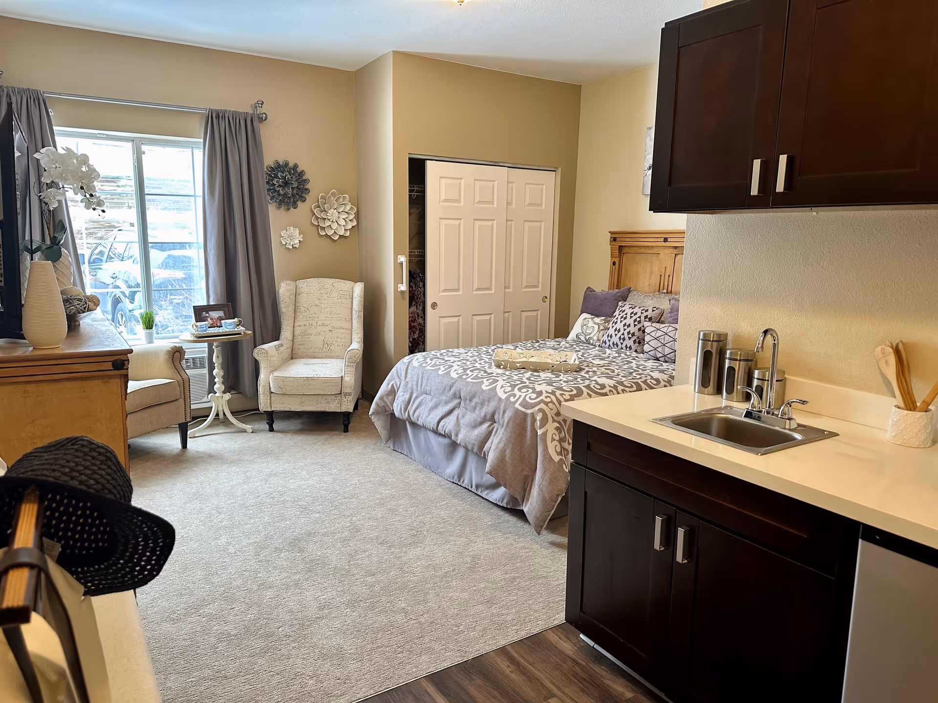 A cozy studio apartment featuring a bed with decorative pillows and a patterned comforter, a small kitchenette with a sink and dark wood cabinets, two upholstered armchairs with a small round table between them near a window with gray curtains, and a closet with sliding doors.