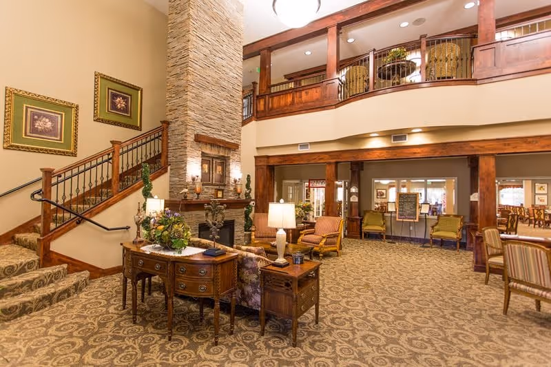 Spacious and warmly decorated living area in a retirement community featuring a stone fireplace, wooden staircase with railing, patterned carpet, multiple chairs and tables with lamps, and framed artwork on the walls. The room has a high ceiling with a balcony overlooking the area.