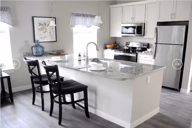 Modern kitchen area with a granite countertop island featuring a sink and two black chairs. The kitchen includes white cabinets, a stainless steel refrigerator, microwave, and stove. There are two windows with patterned valances, a framed botanical print on the wall, and various kitchen items on the counters.