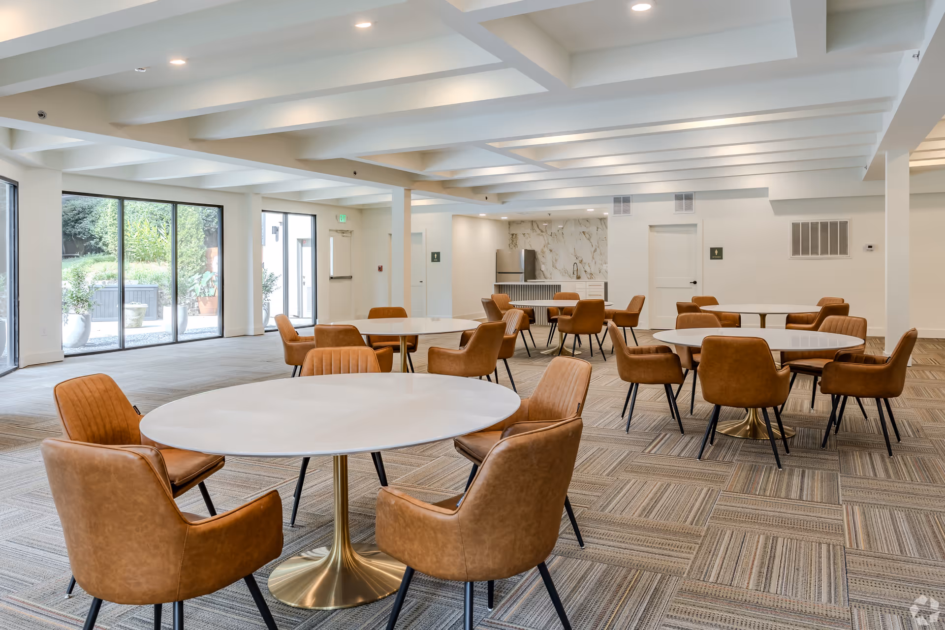 A spacious, modern community room with multiple round white tables surrounded by brown leather chairs. The room has large windows letting in natural light and a small kitchenette area with a marble backsplash in the background. The ceiling features exposed beams with recessed lighting.
