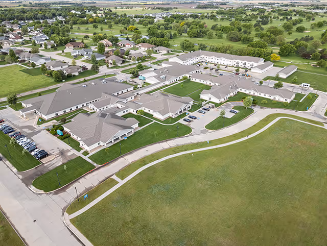 Aerial view of Good Samaritan Society - Grand Island Village facility showing multiple connected buildings with gray roofs, surrounded by green lawns, parking lots with cars, and nearby residential houses and trees.