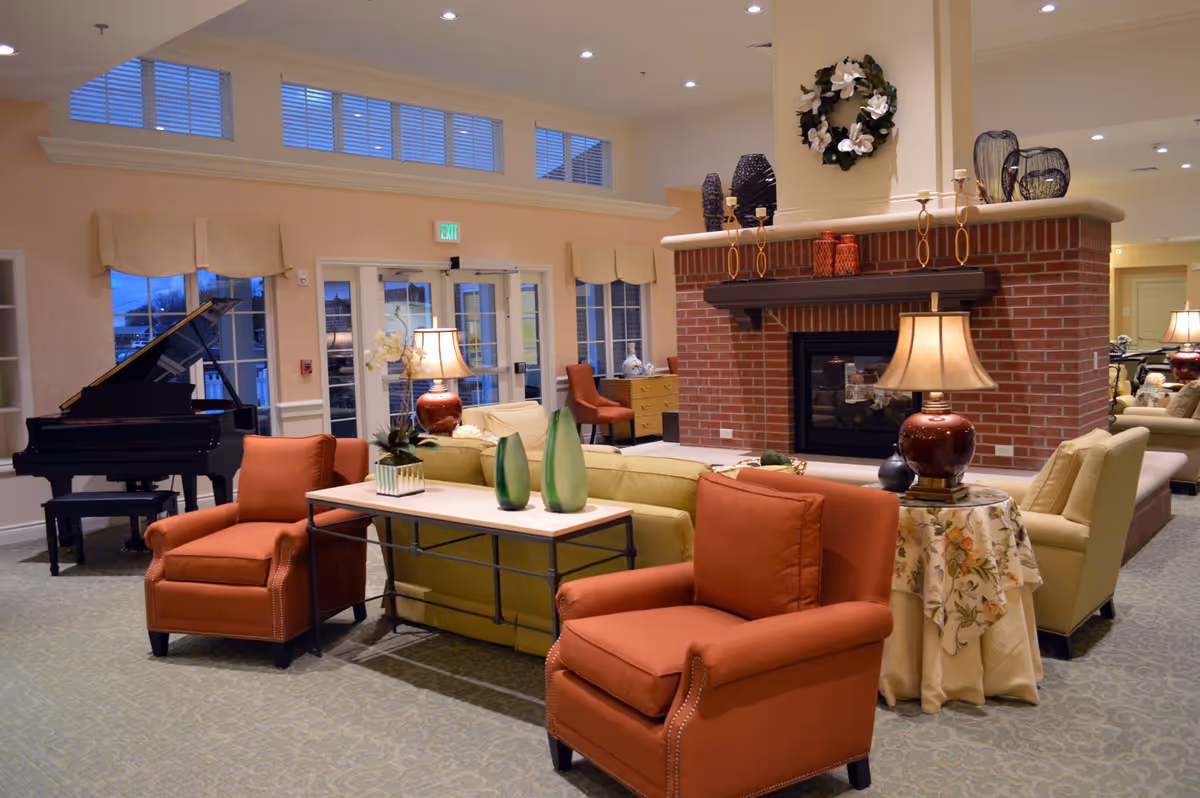 Spacious lounge area with armchairs and sofas arranged around a brick fireplace, decorative accents, lamps, and a grand piano.