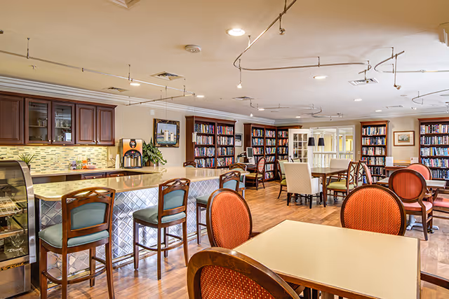 A spacious and well-lit common area with a kitchen counter and bar stools on the left, several tables and chairs arranged for seating, and bookshelves filled with books along the back wall. The room has wooden flooring and ceiling lights, creating a warm and inviting atmosphere.