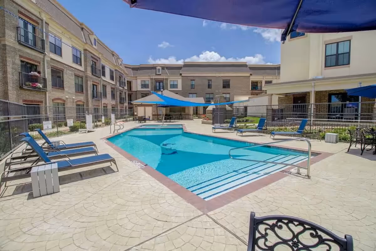 Outdoor swimming pool area at Discovery Village At Castle Hills with blue lounge chairs, blue shade sails, and surrounding multi-story residential buildings under a clear blue sky.