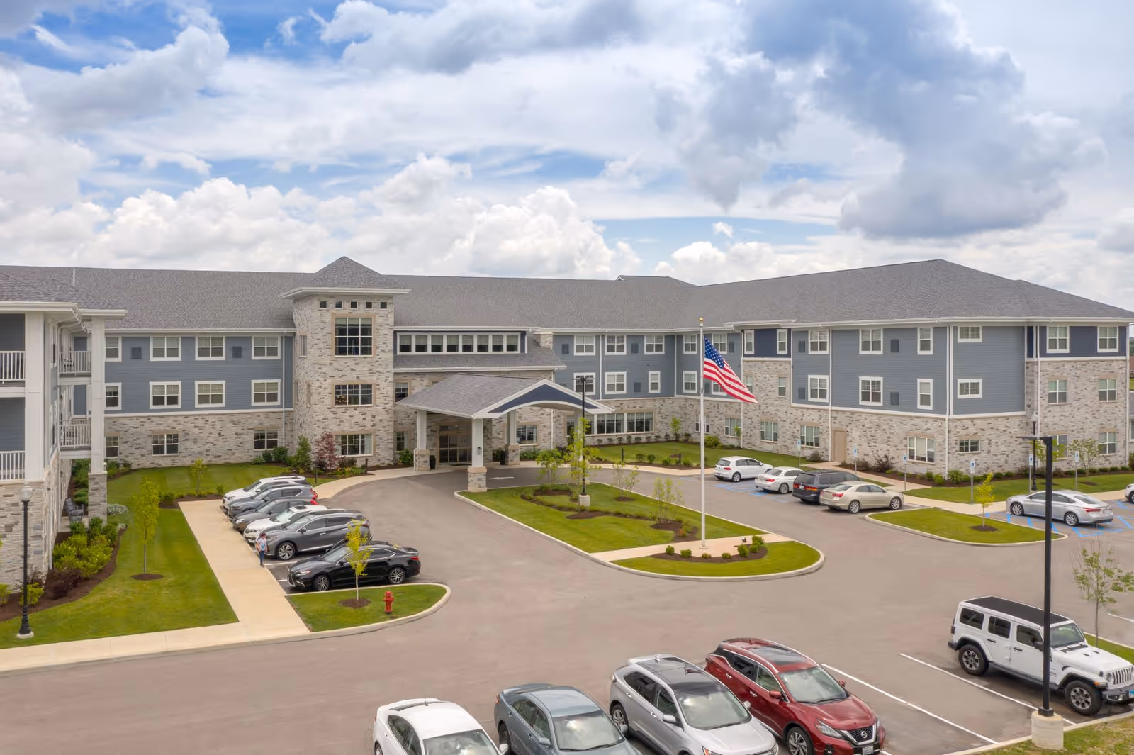 Exterior view of Clarendale of St. Peters senior living facility showing a large three-story building with gray siding and stone accents, a covered entrance, a parking lot with several cars, a flagpole with an American flag, and a partly cloudy sky.