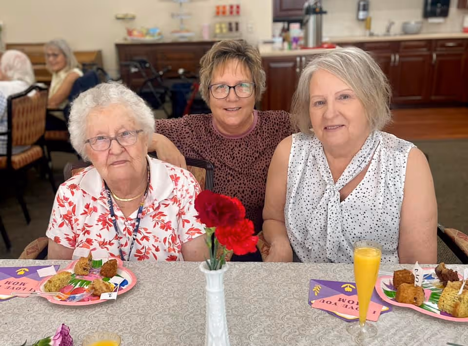 Three elderly women sitting together at a dining table in an assisted living facility. The table is set with plates of food, a glass of orange juice, and a vase with red flowers. The background shows a kitchen area with cabinets and appliances.